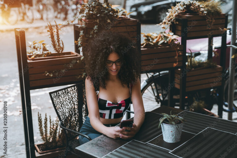 Charming Caucasian female looking at her smartphone screen in surprise while sitting at the table of a cafe outdoors, a cute curly girl with the cell phone in a street bar waiting for her order